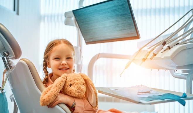 Young girl in dental chair grinning and holding stuffed rabbit toy